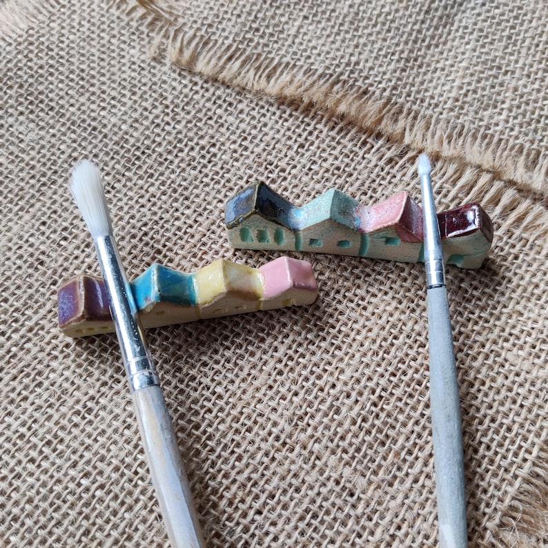 May include: Three ceramic house-shaped brush holders in pink, blue, and brown, with two paintbrushes on a burlap surface.