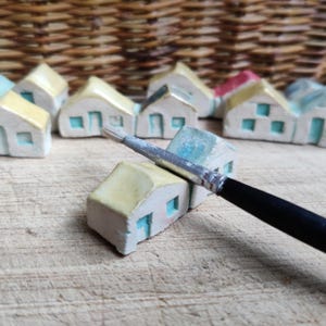 May include: A collection of miniature ceramic houses painted in yellow and blue with a paintbrush resting on top of one of the houses.