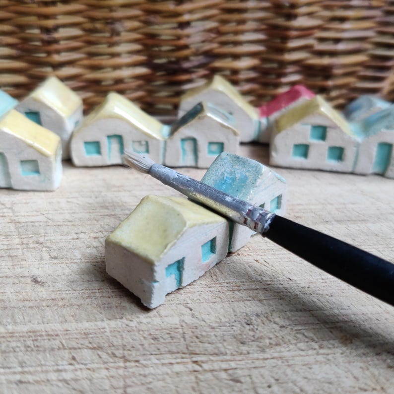 May include: A collection of miniature ceramic houses painted in yellow and blue with a paintbrush resting on top of one of the houses.