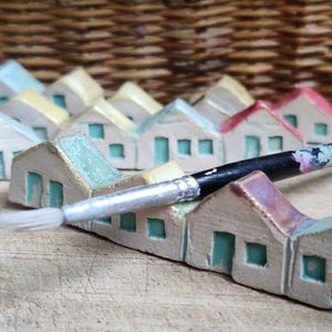 May include: A row of miniature ceramic houses painted in shades of blue, green, and yellow, with a paintbrush resting on top. The houses are arranged in a row on a wooden surface.