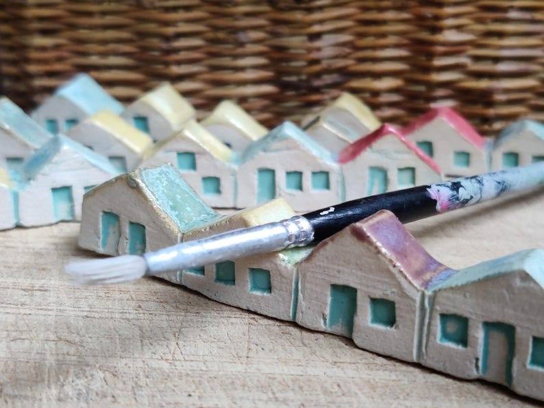May include: A row of miniature ceramic houses painted in shades of blue, green, and yellow, with a paintbrush resting on top. The houses are arranged in a row on a wooden surface.