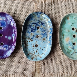 May include: Three ceramic soap dishes in oval shapes with different colour patterns. The dishes are purple with white speckles, blue with white speckles, and green with white speckles.