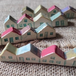 May include: A row of miniature ceramic houses with colorful roofs and painted details. The houses are arranged in a staggered pattern on a burlap background.