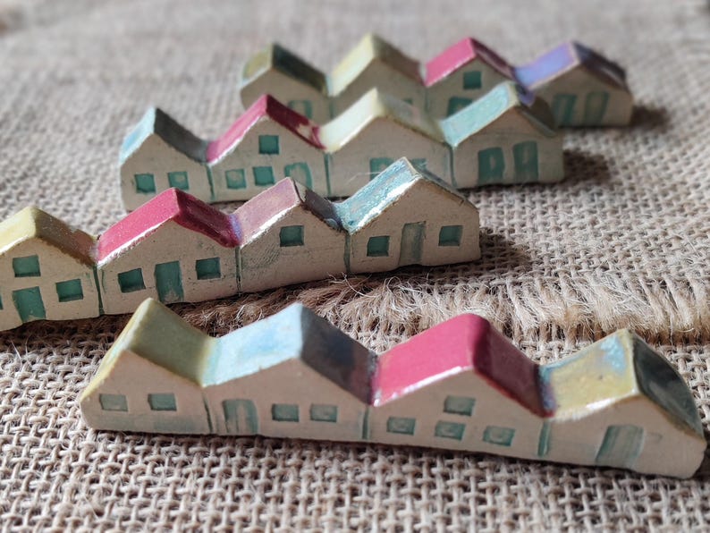May include: A row of miniature ceramic houses with colorful roofs and painted details. The houses are arranged in a staggered pattern on a burlap background.