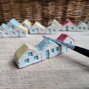 May include: A row of miniature ceramic houses with yellow, blue, and red accents. The houses are arranged in a line on a wooden surface. A paintbrush is lying on the surface next to the houses.