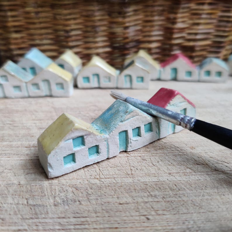 May include: A row of miniature ceramic houses with yellow, blue, and red accents. The houses are arranged in a line on a wooden surface. A paintbrush is lying on the surface next to the houses.
