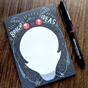 Puede incluir: Un bloc de notas negro con una ilustración de bombilla blanca y el texto "BRIGHT IDEAS". La bombilla tiene ojos rojos y un espacio en blanco para escribir.