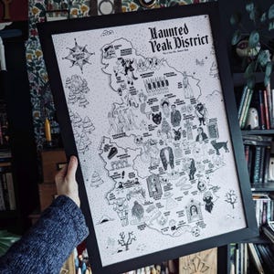 May include: A black and white illustrated map of the Peak District in England, titled "Haunted Peak District." The map features various spooky and mythical creatures, including ghosts, witches, and monsters. The map is framed in a black frame.