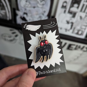 Peut inclure: Épingle à nourrice en forme de papillon de nuit noir avec des yeux rouges, sur fond d'éclat blanc. La carte noire porte un dessin de fantôme et le texte "boo!". Le texte "@ThisisSianEllis" est également présent.