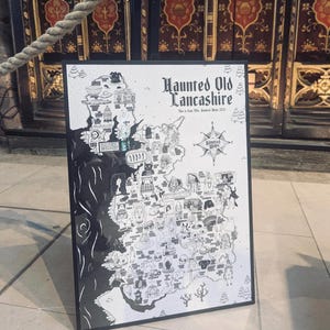 May include: A black and white framed map titled Haunted Old Lancashire, featuring various illustrated icons and text labels across the region, a compass rose, and a decorative border, presented in a vertical orientation on a display stand.