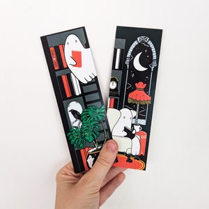 K&ouml;nnte beinhalten: Zwei schwarze Lesezeichen mit Illustrationen von Geistern, die B&uuml;cher lesen. Ein Lesezeichen zeigt einen Geist, der in einem B&uuml;cherregal mit einem Raben und einer Monstera-Pflanze ein Buch liest. Das andere Lesezeichen zeigt einen Geist, der in einem Stuhl mit einem Halbmond und einem roten Lampenschirm im Hintergrund ein Buch liest. Der Text "GHOST STORIES" ist auf dem B&uuml;cherregal sichtbar.