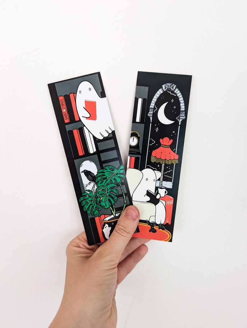 K&ouml;nnte beinhalten: Zwei schwarze Lesezeichen mit Illustrationen von Geistern, die B&uuml;cher lesen. Ein Lesezeichen zeigt einen Geist, der in einem B&uuml;cherregal mit einem Raben und einer Monstera-Pflanze ein Buch liest. Das andere Lesezeichen zeigt einen Geist, der in einem Stuhl mit einem Halbmond und einem roten Lampenschirm im Hintergrund ein Buch liest. Der Text "GHOST STORIES" ist auf dem B&uuml;cherregal sichtbar.