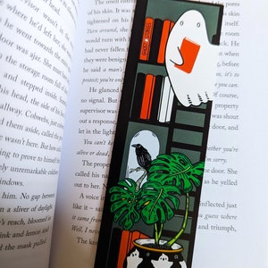 Puede incluir: Un marcapáginas negro y verde con un fantasma blanco que sostiene un libro rojo. El fantasma está de pie en una estantería con las palabras "Ghost Stories" impresas en el lateral. El marcapáginas también presenta un cuervo negro posado en una rama de una planta de monstera verde en una maceta negra con fantasmas blancos y estrellas. La maceta está sobre una superficie marrón.