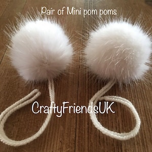 May include: Two fluffy white mini pom poms with white cords. The image includes the text "Pair of Mini pom poms" and "CraftyFriendsUK".