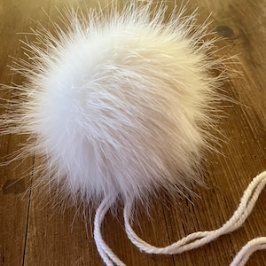 May include: A large, fluffy white fur pompom with long, soft strands. Two white strings are attached, possibly for securing the pompom to a hat or garment. The pompom is displayed against a wooden surface.