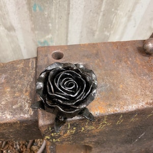 Puede incluir: Escultura artesanal de una rosa de metal negro. La rosa presenta pétalos superpuestos y detallados. Se exhibe sobre una superficie metálica desgastada, resaltando la textura y la artesanía.