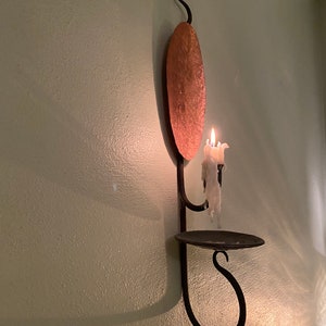 May include: A black metal wall-mounted candle holder with a copper plate and a lit white candle. The candle holder has a shelf and a hook.