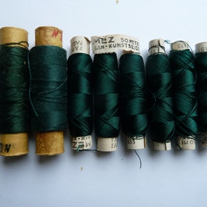 May include: Eight spools of dark green thread, each wrapped in paper with text that reads "MEZ 50 METER LAN-KUNSTSEIDE" and "FAR", "NO", "ARB", "1453", "BW OS", "03", and "153".