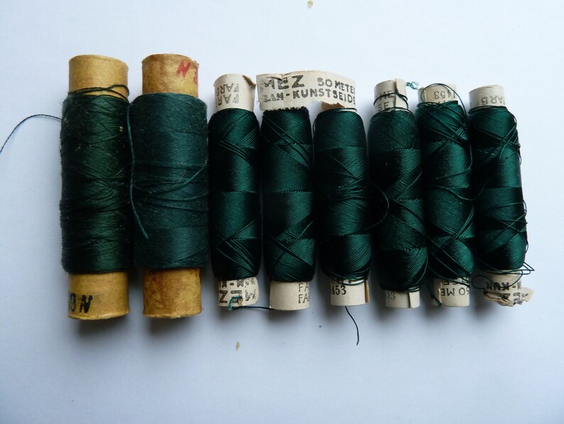 May include: Eight spools of dark green thread, each wrapped in paper with text that reads "MEZ 50 METER LAN-KUNSTSEIDE" and "FAR", "NO", "ARB", "1453", "BW OS", "03", and "153".