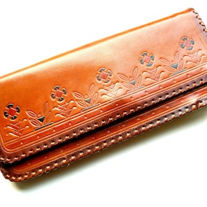 Könnte beinhalten: Eine braune Leder-Clutch mit einem dekorativen, gesteppten Rand und einem geprägten Blumenmuster in Braun-, Rot- und Schwarztönen.