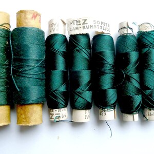 May include: Eight spools of dark green thread, each wrapped in paper with printed text, including "50 METE LAN-KUNST SEIDE" and "FA 153".