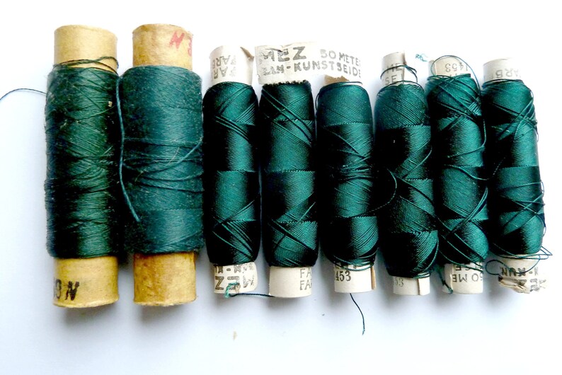May include: Eight spools of dark green thread, each wrapped in paper with printed text, including "50 METE LAN-KUNST SEIDE" and "FA 153".