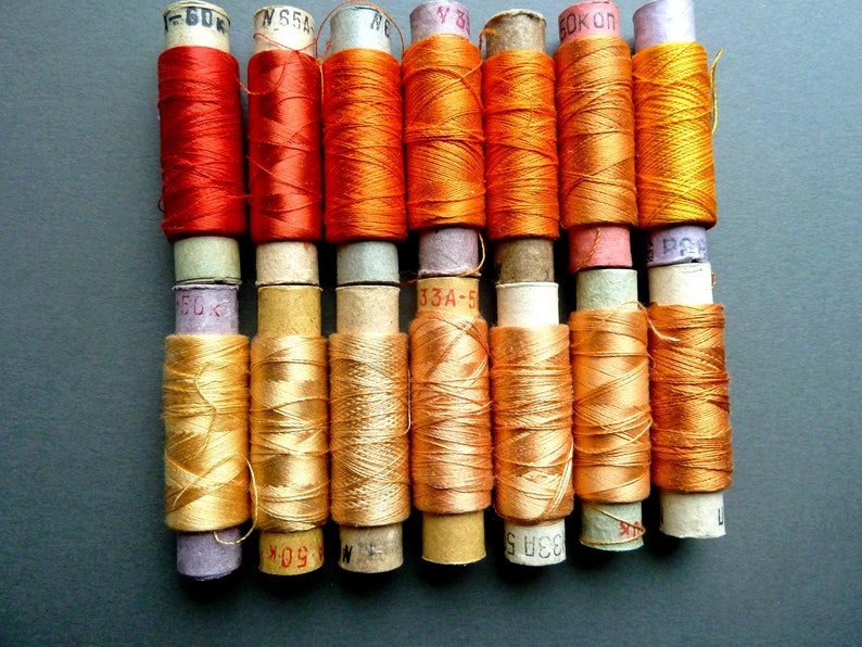 May include: A collection of 10 spools of thread, with a mix of orange and gold colors. The spools are labeled with numbers and letters.