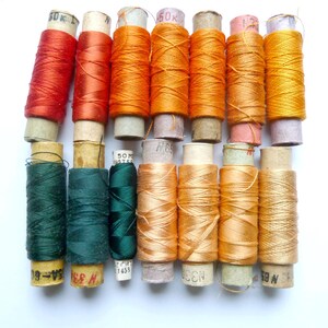 May include: A collection of 12 spools of thread in various shades of orange, green, and red. The spools are arranged in two rows, with the top row containing six spools and the bottom row containing six spools.