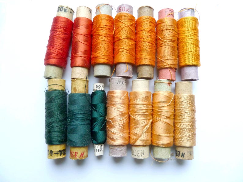 May include: A collection of 12 spools of thread in various shades of orange, green, and red. The spools are arranged in two rows, with the top row containing six spools and the bottom row containing six spools.