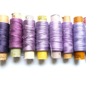May include: A collection of nine spools of thread in various shades of purple, lavender, and gray. The spools are wrapped in paper with labels that include numbers and letters.