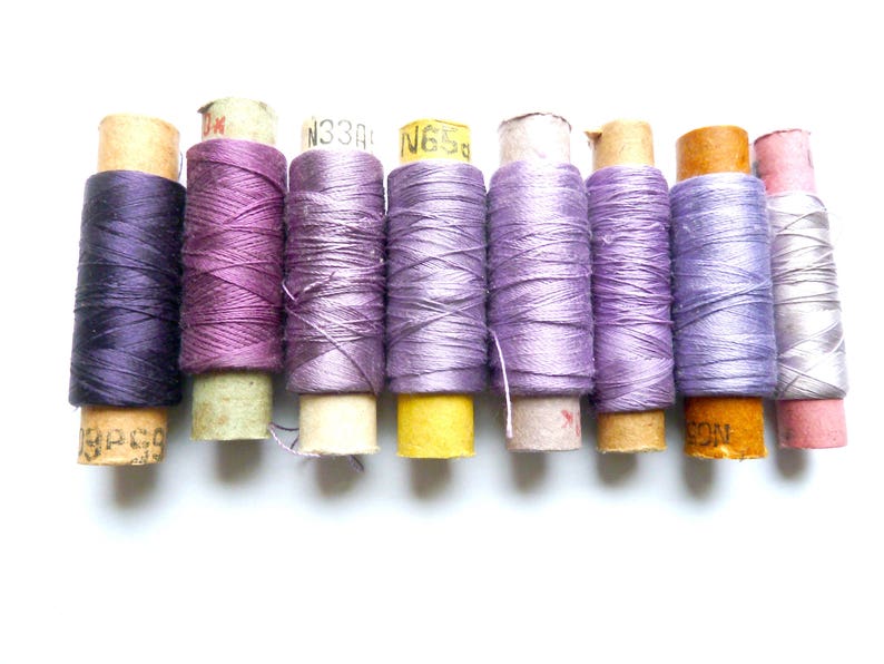 May include: A collection of nine spools of thread in various shades of purple, lavender, and gray. The spools are wrapped in paper with labels that include numbers and letters.