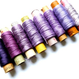 May include: A collection of spools of thread in various shades of purple, lavender, and pink. The spools are wrapped in paper and have labels with numbers and letters.