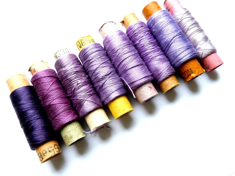 May include: A collection of spools of thread in various shades of purple, lavender, and pink. The spools are wrapped in paper and have labels with numbers and letters.