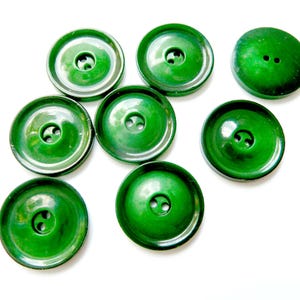 May include: Eight glossy, round, emerald green buttons are displayed. Each button has either two or one hole for sewing. The buttons are set against a white backdrop, highlighting their vibrant colour and design.