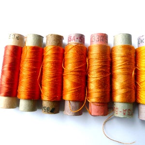 May include: A close-up of seven spools of orange thread. The spools are wrapped in brown paper and labeled with numbers and letters.