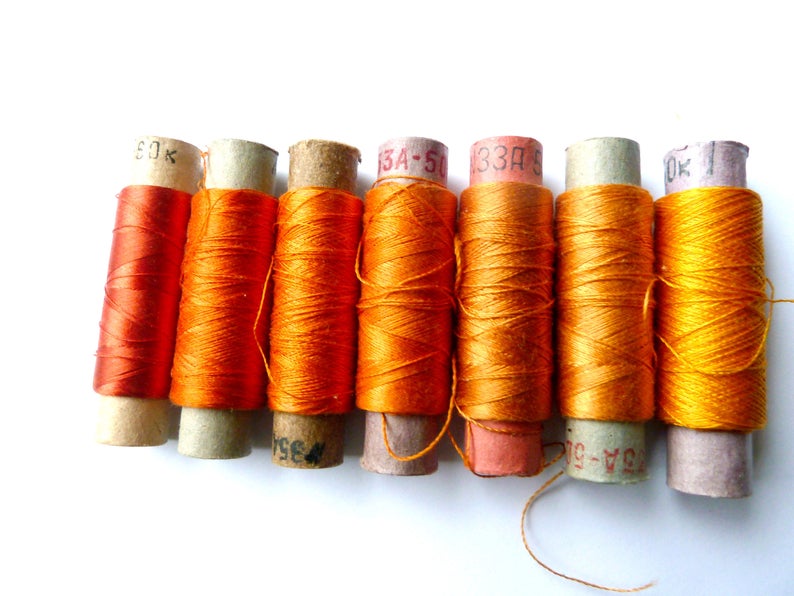 May include: A close-up of seven spools of orange thread. The spools are wrapped in brown paper and labeled with numbers and letters.