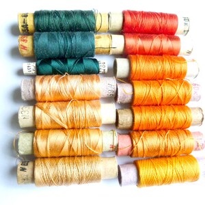 May include: A collection of spools of thread in various shades of orange, green, and red. The spools are wrapped in paper with printed labels and numbers.