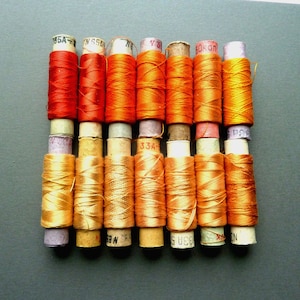 May include: A collection of 12 spools of thread in various shades of orange and red. The spools are labeled with numbers, including "195A", "N 65A", "76", "Y 3", "60K", and "33A".