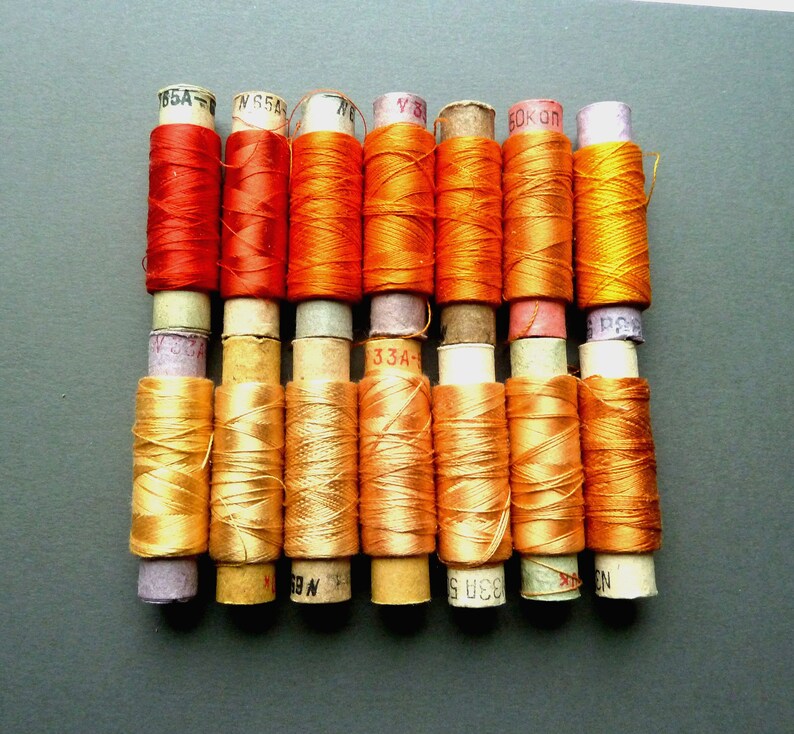 May include: A collection of 12 spools of thread in various shades of orange and red. The spools are labeled with numbers, including "195A", "N 65A", "76", "Y 3", "60K", and "33A".
