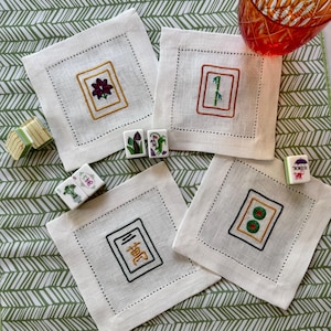 May include: Four square white linen coasters with embroidered Mahjong tile designs. Each coaster features a different tile symbol in a gold or brown frame. Mahjong tiles and a decorative glass are also present.
