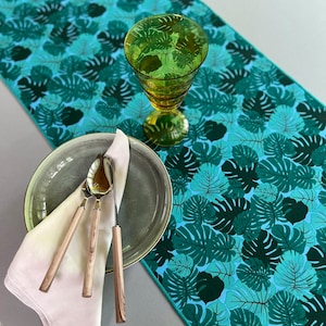 May include: A table setting featuring a teal and green monstera leaf patterned table runner. A green glass, a gray plate with silverware, and a white napkin are arranged on the runner. The runner adds a tropical touch to the dining table.