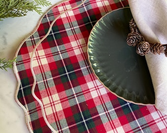 May include: Two red and green plaid placemats with scalloped edges and gold trim. A dark green plate and a beige napkin are on top of the placemats.