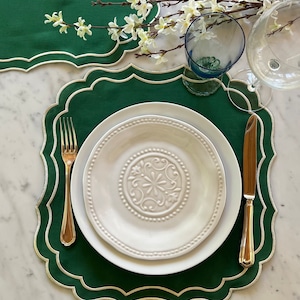 Può includere: Un piatto bianco da cena con un design decorativo si trova su un sottopiatto verde con bordo a festoni. Una forchetta e un coltello dorati sono posizionati su entrambi i lati del piatto.