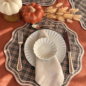 May include: A scalloped-edge placemat with a brown and black plaid pattern, featuring a white plate, a small fluted bowl, a beige linen napkin, a silver fork, a silver knife, a silver spoon, a white pumpkin, an orange pumpkin, and dried grass.