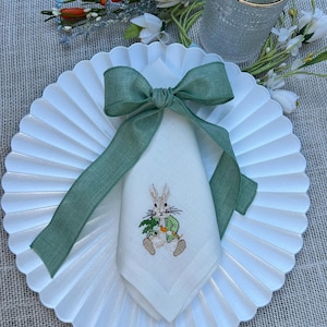 Peut inclure: Une serviette en lin blanc avec un lapin brodé tenant une carotte. La serviette est nouée avec un ruban vert et placée sur une assiette blanche à festons.