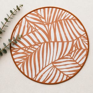 May include: A circular placemat featuring a white background with a repeating pattern of terracotta-colored palm leaves. The placemat has a solid terracotta-colored border around the edge. A sprig of green eucalyptus is placed to the left side.