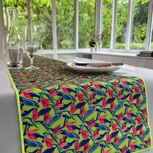 May include: A green table runner with a repeating pattern of pink and blue tropical flowers. The runner has a lime green border.