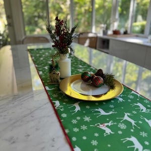 May include: A festive Christmas table setting featuring a green runner with white reindeer and snowflake patterns. A gold-rimmed plate holds ornaments and pinecones. A white vase with red berries and greenery adds a seasonal touch.