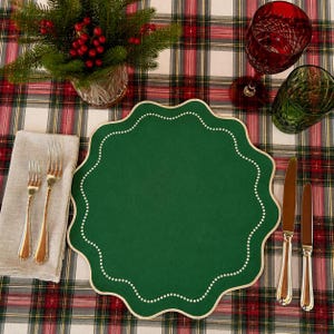 May include: A round, scalloped-edge dark green placemat featuring a delicate white dotted border. The placemat is set on a red, green, and white plaid tablecloth, accompanied by two silver forks, two silver knives, a red glass, and a green glass.