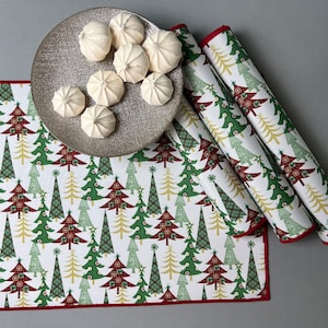 Puede incluir: Un juego de tres caminos de mesa blancos con borde rojo y un patrón repetido de árboles de Navidad verdes. Los caminos de mesa están doblados y dispuestos sobre una superficie gris. Un plato plateado con merengues blancas está en el fondo.